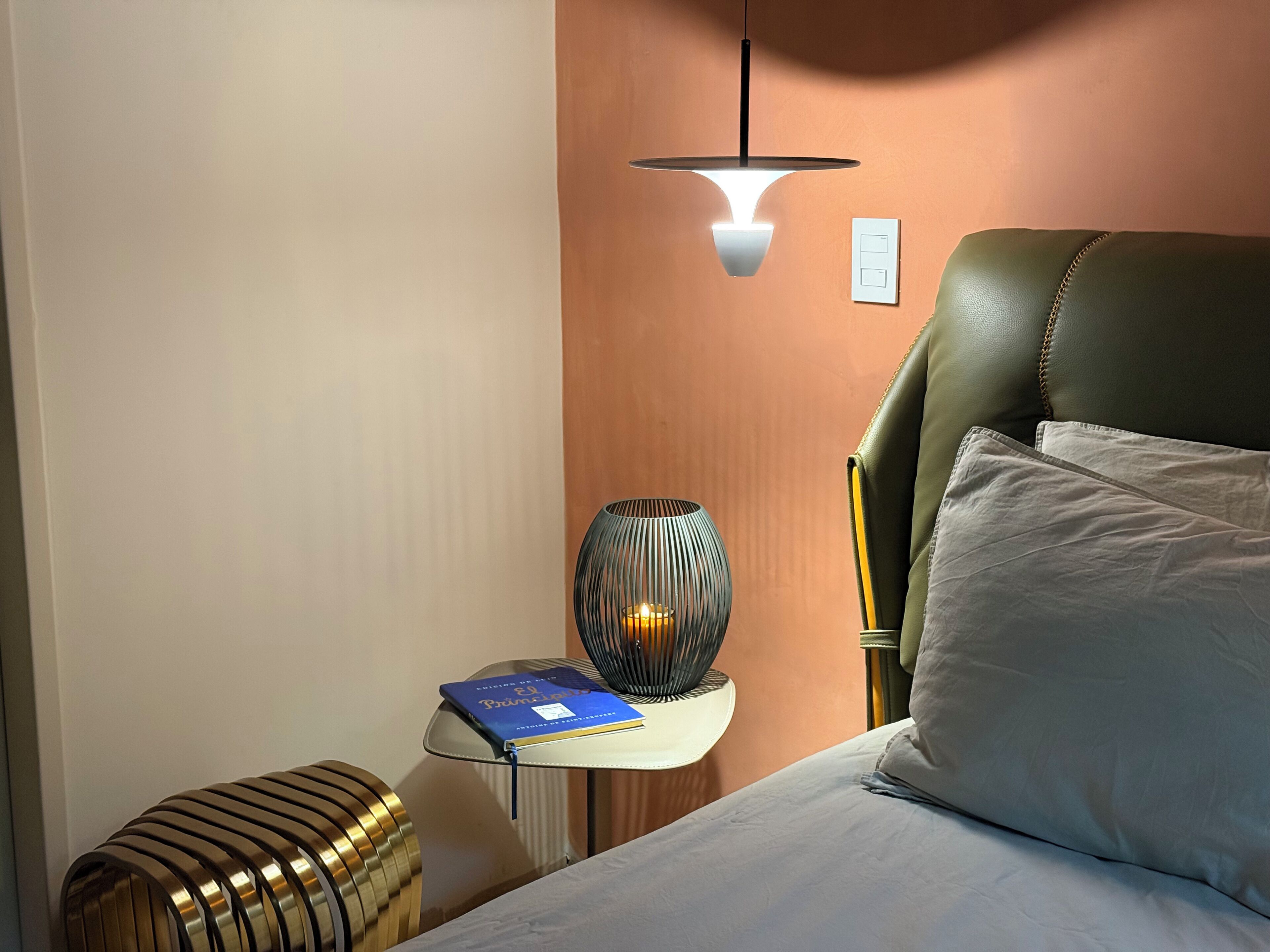 A neatly made bed with a pillow, a bedside table with a book and a lamp, and a wall-mounted light fixture.