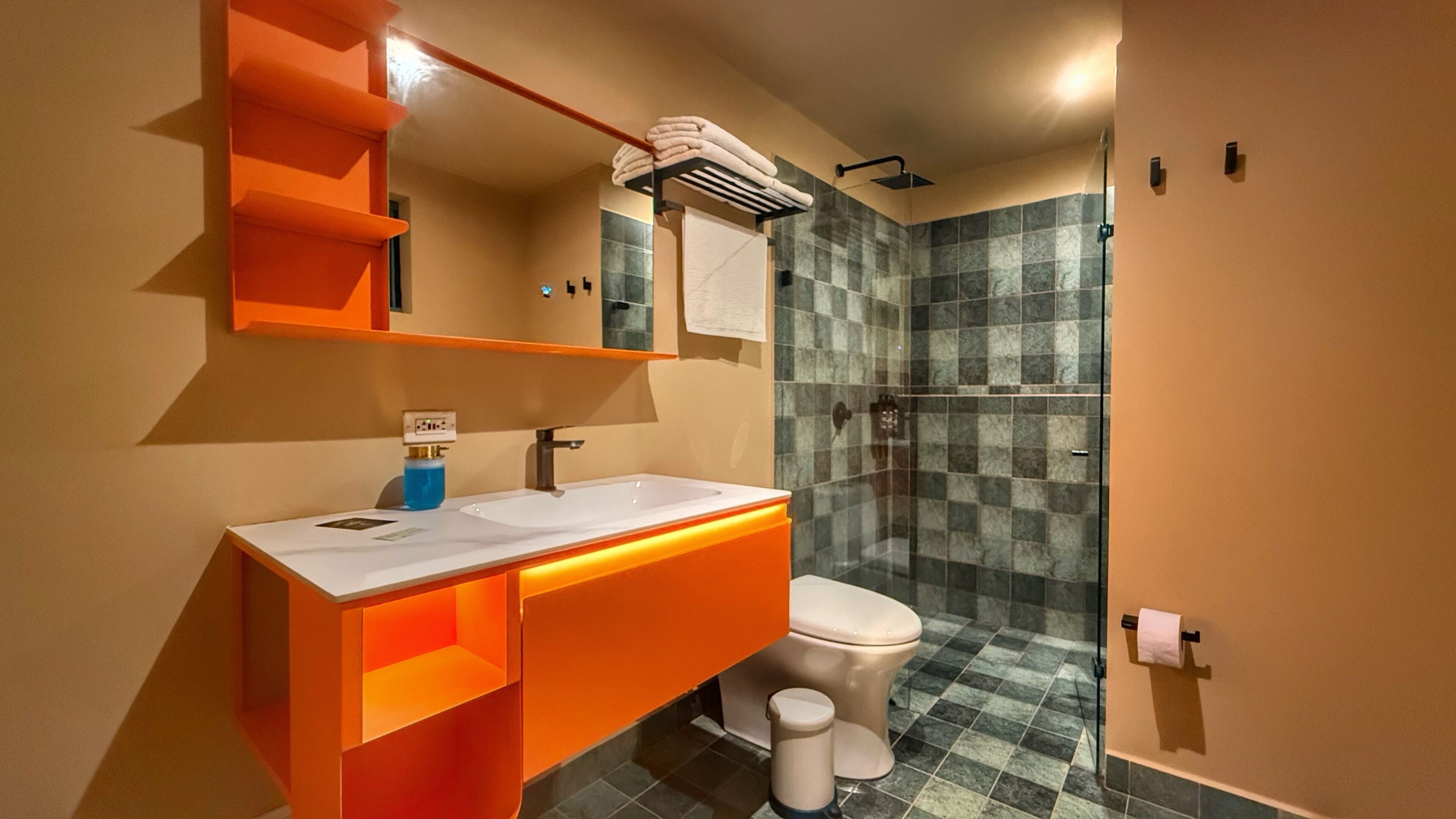 Un baño con lavamanos blanco, tocador naranja, cabina de ducha de vidrio y suelo de baldosas.