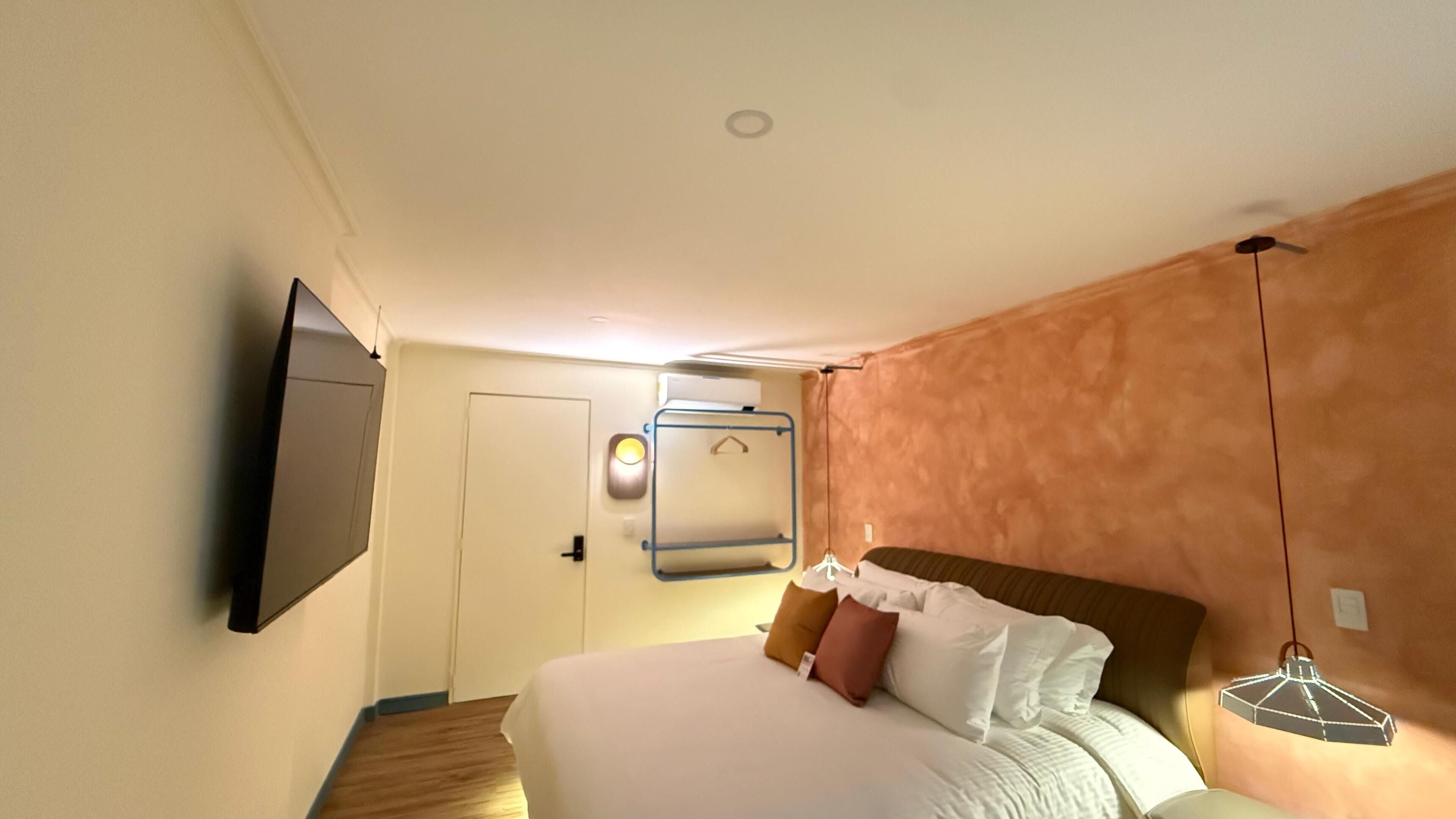 Habitación de hotel con cama, televisor montado en la pared, puerta y lámpara colgante.