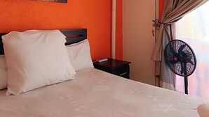 Une chambre d’hôtel avec un lit, un tableau représentant des fleurs, un ventilateur fixé au mur et une fenêtre avec des rideaux.