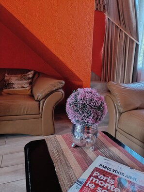 Ein Wohnzimmer mit einer braunen Couch, einem kleinen Tisch mit einer Zeitschrift und einer Vase mit einem rosafarbenen, kugelförmigen Gegenstand.