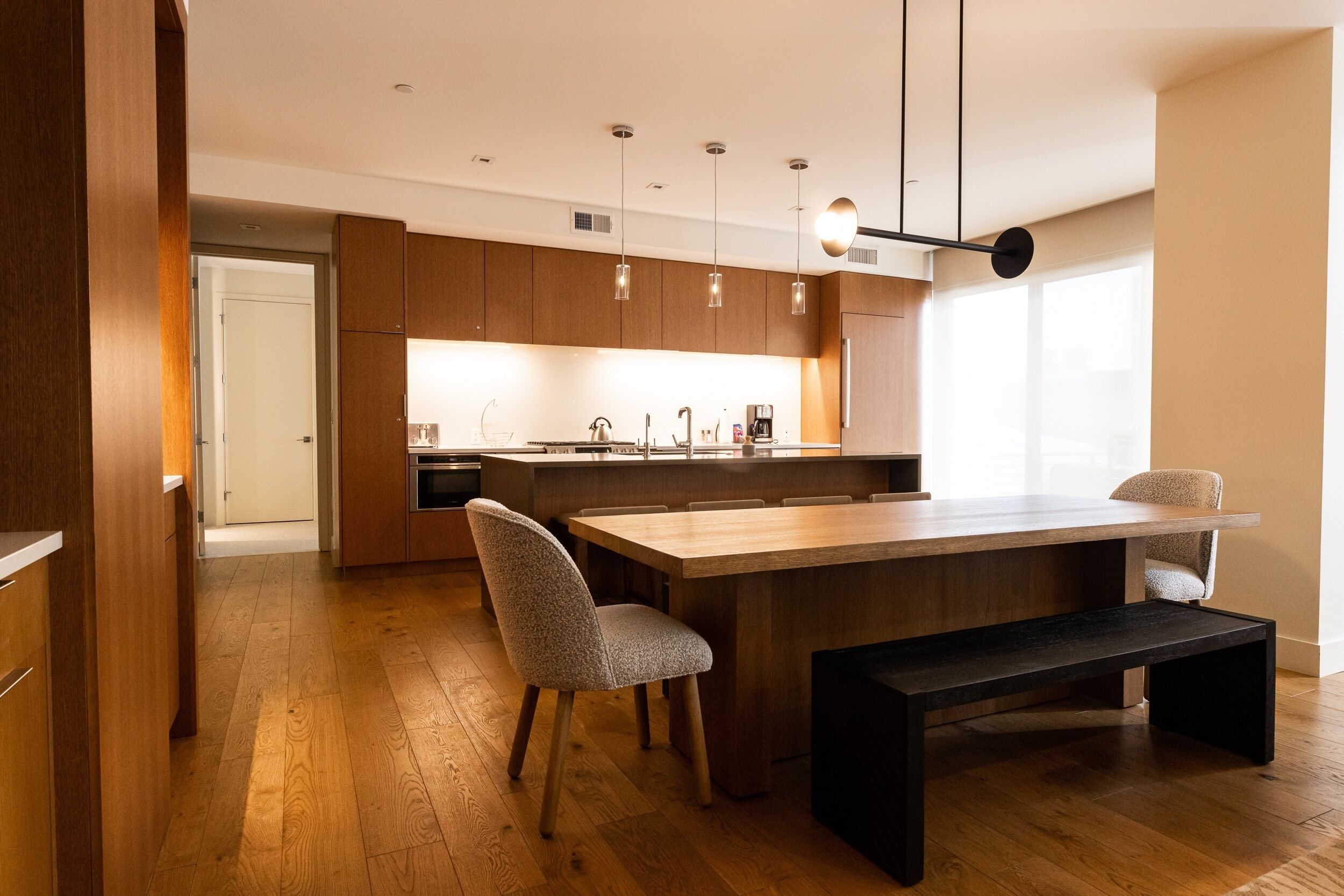 A modern kitchen with wooden cabinets, a large island, pendant lights, and a bench.