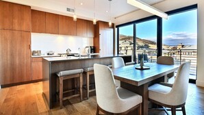 Una cocina moderna con armarios de madera, una isla central y taburetes de bar. Cuenta con ventanas amplias que ofrecen vistas a un paisaje costero.