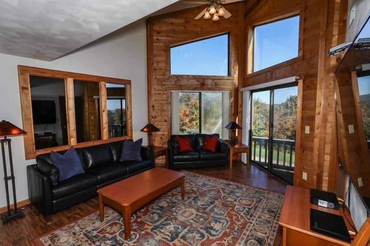 A spacious living room with a black leather sofa, a wooden coffee table, and a patterned rug. There are large windows offering a view of the outdoors.