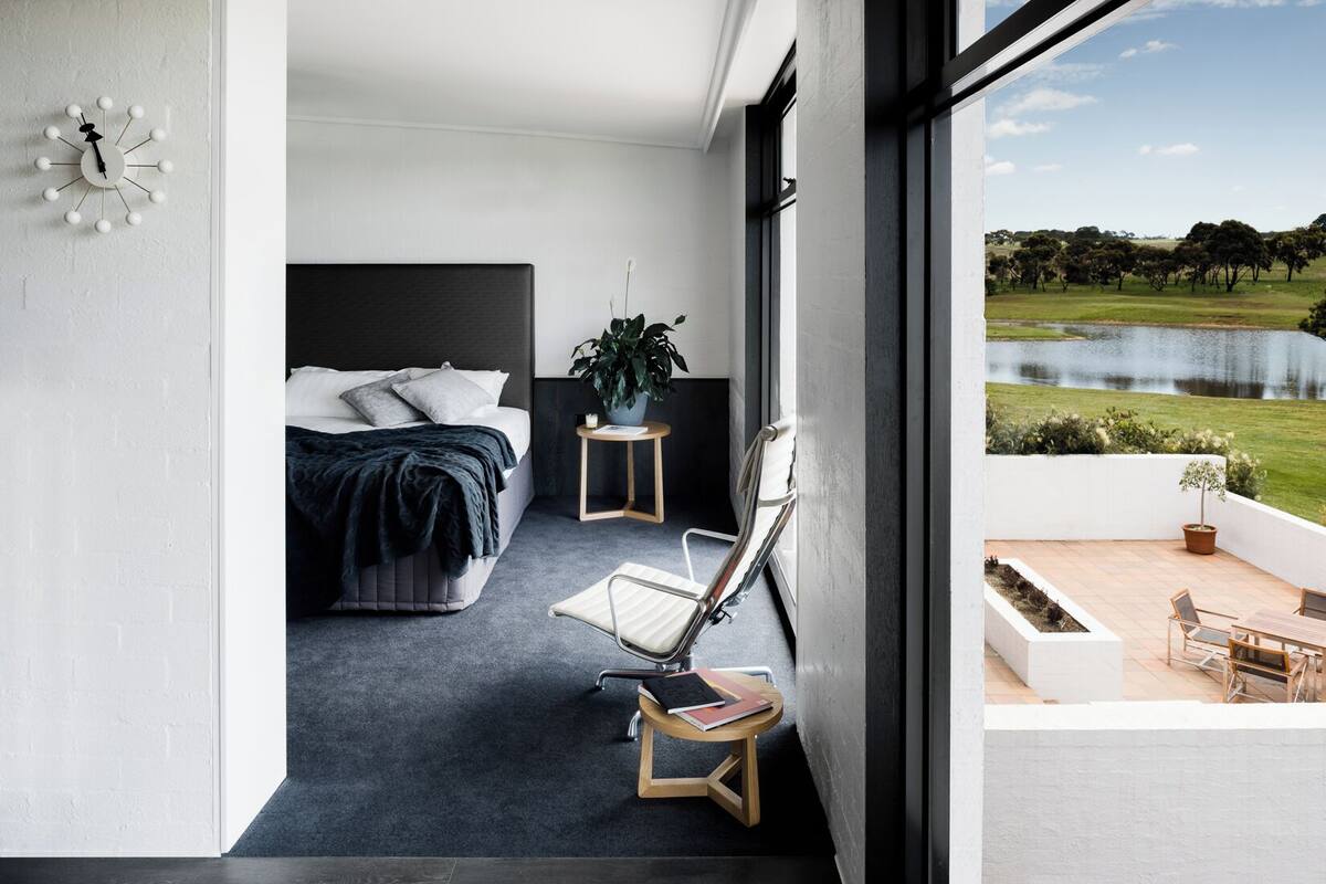 A modern hotel room with a bed, a small side table with a plant, a chair, and a view of a lake and greenery outside.