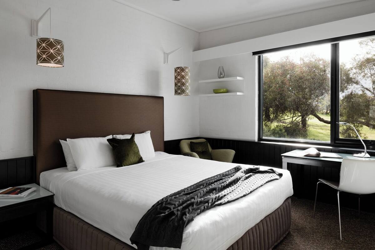 A modern hotel room with a large bed, a desk, and a view of trees through the window.