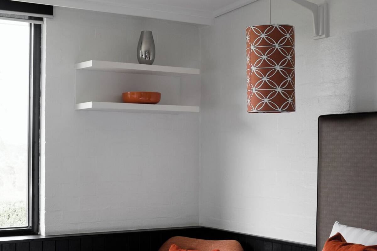 A modern hotel room with a bed, a chair, a shelf with decorative items, and a hanging light fixture.