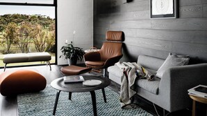 A modern living room with a grey sofa, a brown leather chair, a round coffee table, and a large window offering a view of trees. - Waurn Ponds Estate (Waurn Ponds)