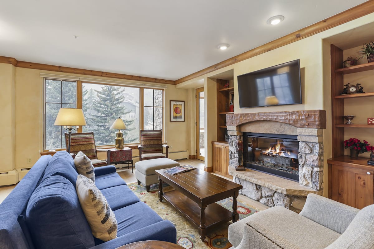 A living room with a stone fireplace, a large TV mounted above it, and a comfortable seating area with a sofa and armchairs.