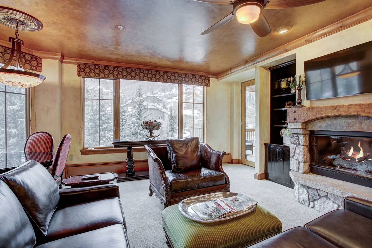 A cozy living room with leather furniture, a fireplace, and large windows overlooking a snowy landscape.