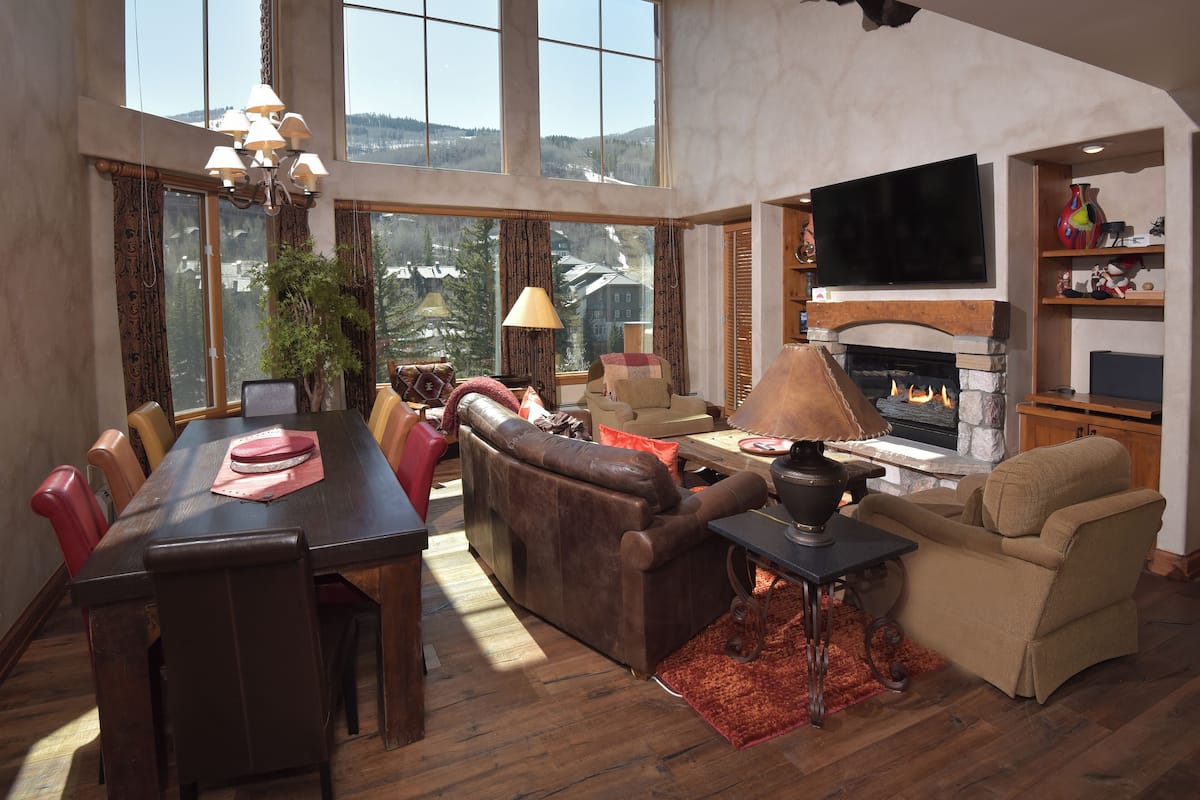 A spacious living room with a fireplace, a dining area with a table and chairs, and a large window offering a view of the outdoors.