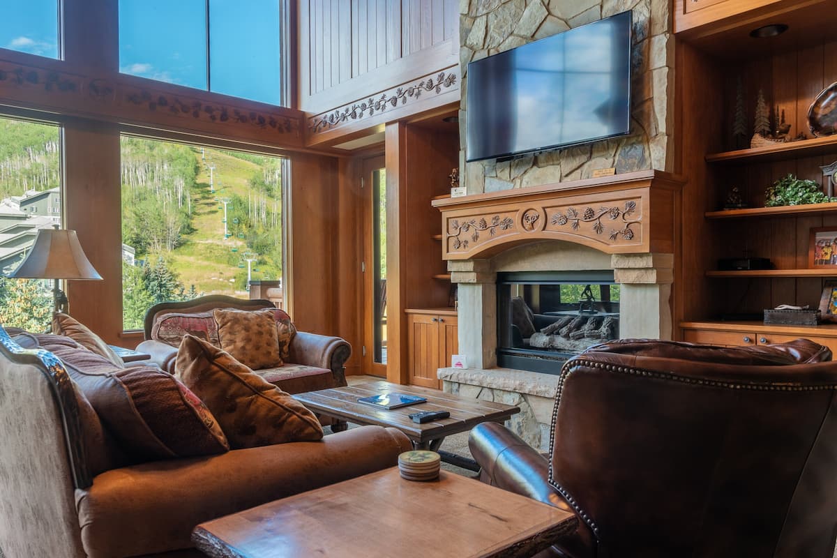 A cozy living room with a fireplace, leather furniture, and a large window offering a view of a forested area.