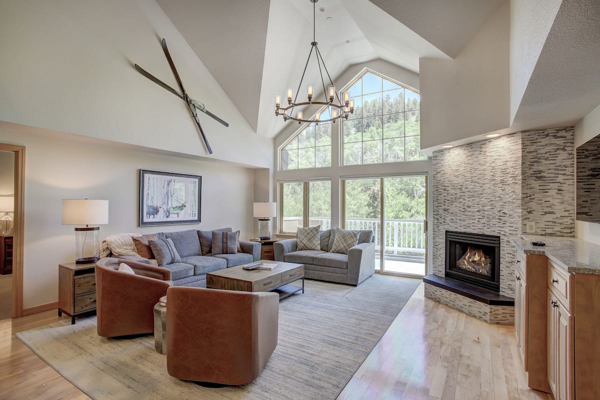 A spacious living room with a fireplace, large windows, and a high ceiling.