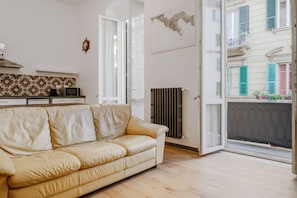 Une pièce avec un canapé beige, un coin cuisine, un radiateur et une fenêtre donnant sur des immeubles.