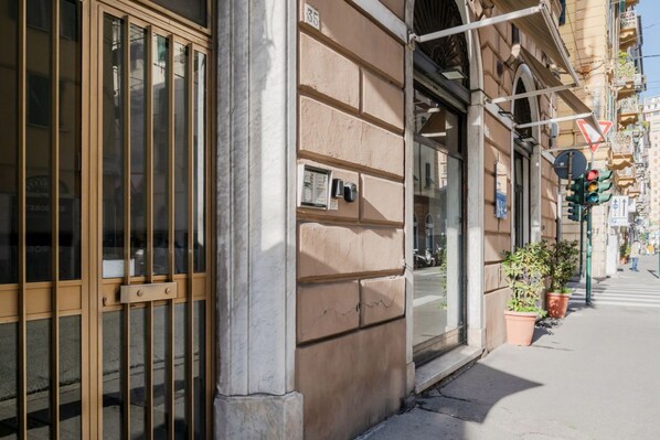 Une façade d’immeuble avec de grandes fenêtres, un portail en métal et une rue bordée de plantes en pot.