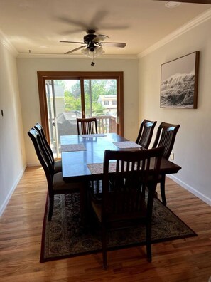 Une salle à manger avec une table et des chaises, un ventilateur de plafond, une grande fenêtre et un tableau encadré au mur.