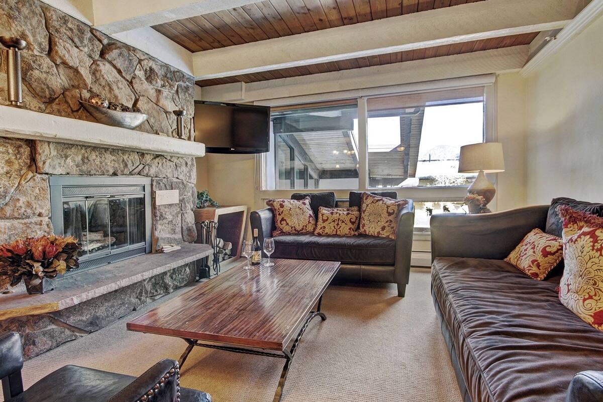 A cozy living room with a stone fireplace, leather sofas, a wooden coffee table, and a large window offering a view of the outdoors.