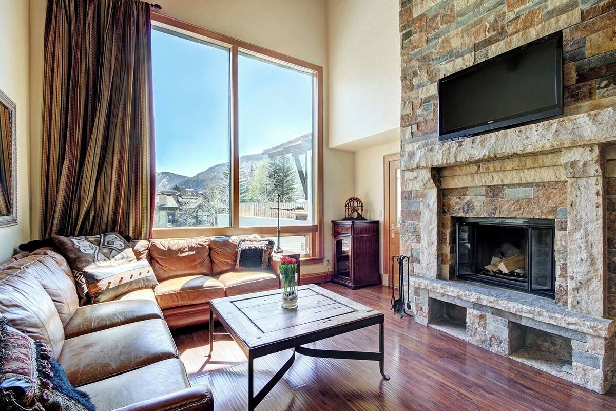 A living room with a stone fireplace, leather sofa, and a large window with a mountain view.