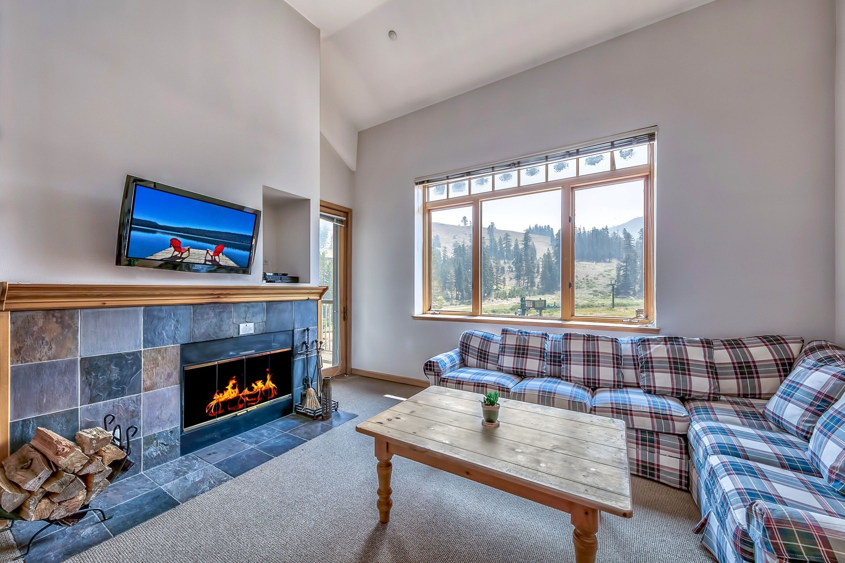 Ein Wohnzimmer mit Kamin, Fernseher, einer Couch und einem großen Fenster mit Blick ins Freie.