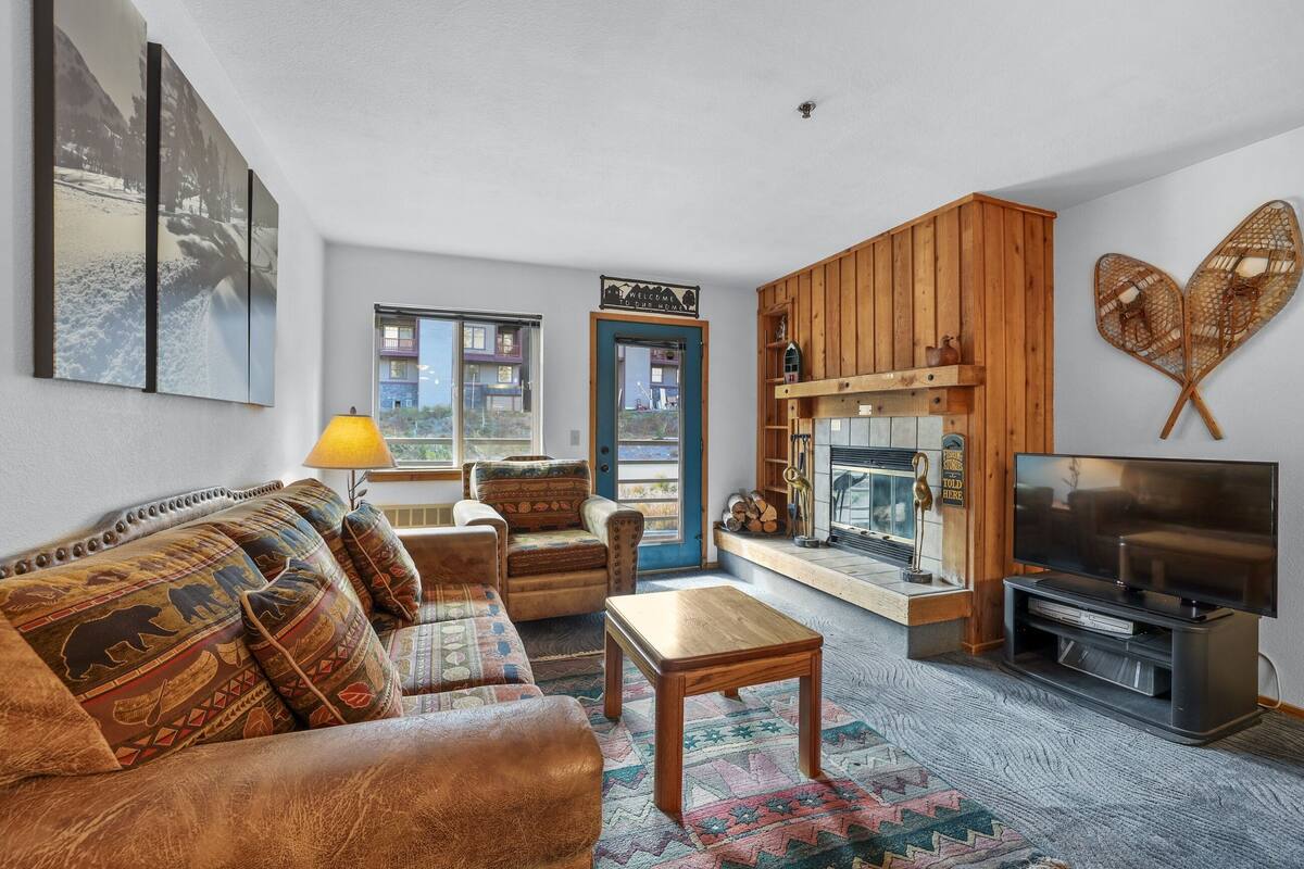 Una sala de estar acogedora con sofá, mesa de centro y mueble para la televisión. Se observa panelado de madera y una chimenea.
