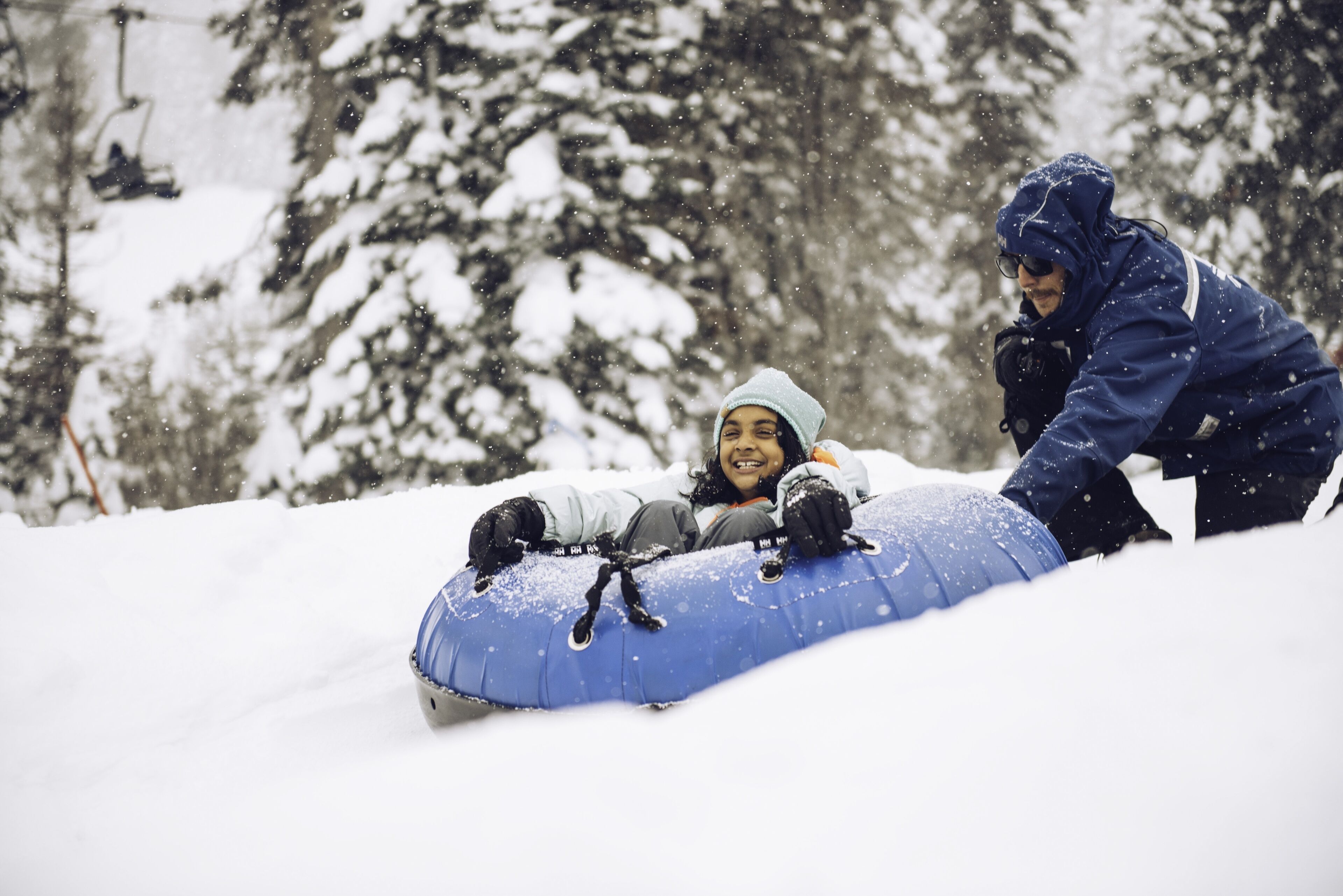 A person lying on a blue inflatable tube in the snow, with another person standing nearby.