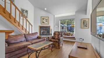 Amplia sala de estar con una escalera de madera, chimenea, sofá, sillón estampado y un ventanal con vistas a casas.