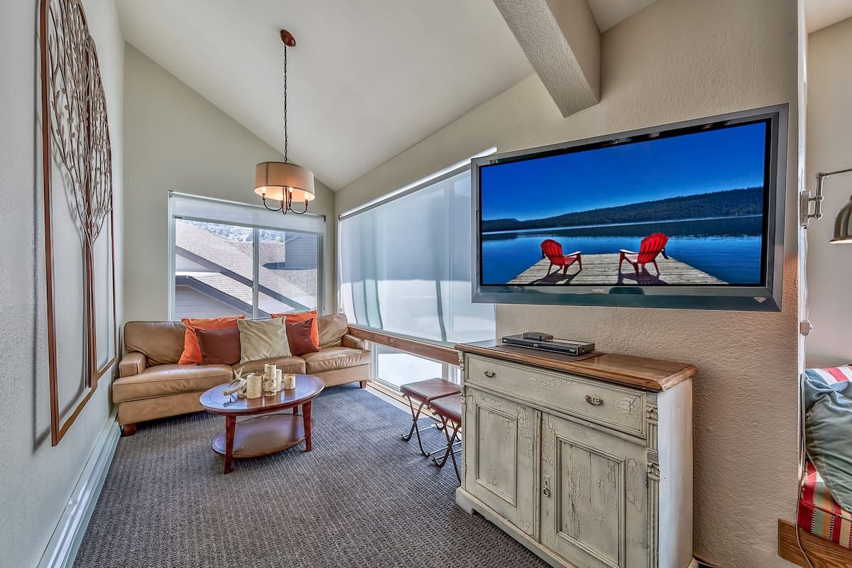 Una sala de estar con televisor grande, un sofá, una mesa de centro y vistas a un muelle con sillas.