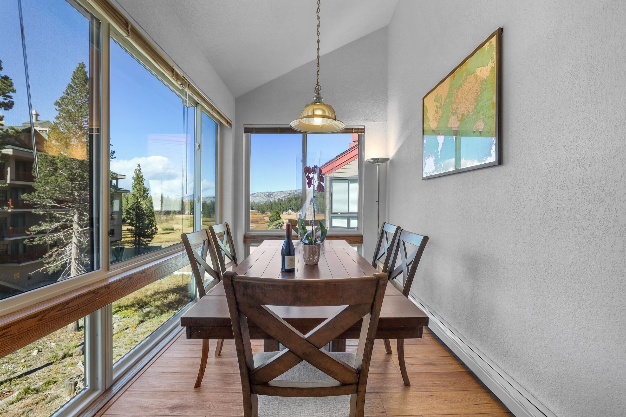 A dining area with a wooden table and chairs, large windows, and a hanging light fixture.