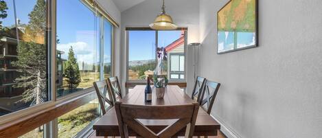 A dining area with a wooden table and chairs, large windows, and a hanging light fixture.