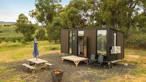 A small, dark-colored cabin with a sliding door, a small patio area with a table and chairs, and a fire pit. - Olive Sanctuary by Tiny Away (Taggerty)