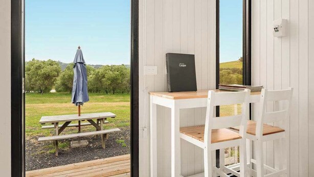 A room with a white desk and chair, a laptop, and a view of an outdoor picnic area with a table and umbrella. - Olive Sanctuary by Tiny Away (Taggerty)