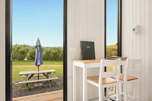 A room with a white desk and chair, a laptop, and a view of an outdoor picnic area with a table and umbrella. - Olive Sanctuary by Tiny Away (Taggerty)