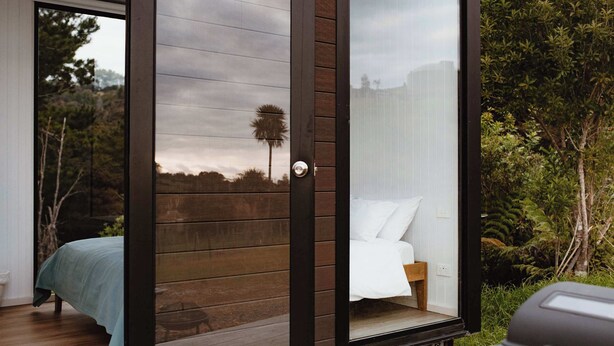 Un dormitorio moderno con un amplio ventanal corredero que da a un espacio exterior, una cama con ropa de cama blanca y vistas al follaje circundante.