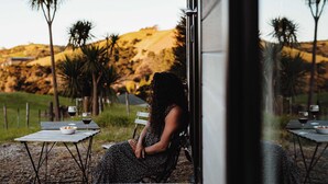 Una mujer sentada a una mesa con un cuenco de fruta y dos copas de vino tinto, contemplando un paisaje pintoresco con colinas y palmeras.