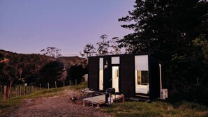 Una cabaña moderna con un amplio ventanal, un pequeño porche y un espacio exterior con mobiliario.