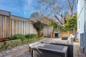 Un patio trasero con terraza de madera, mobiliario exterior, una parrilla y un árbol grande.