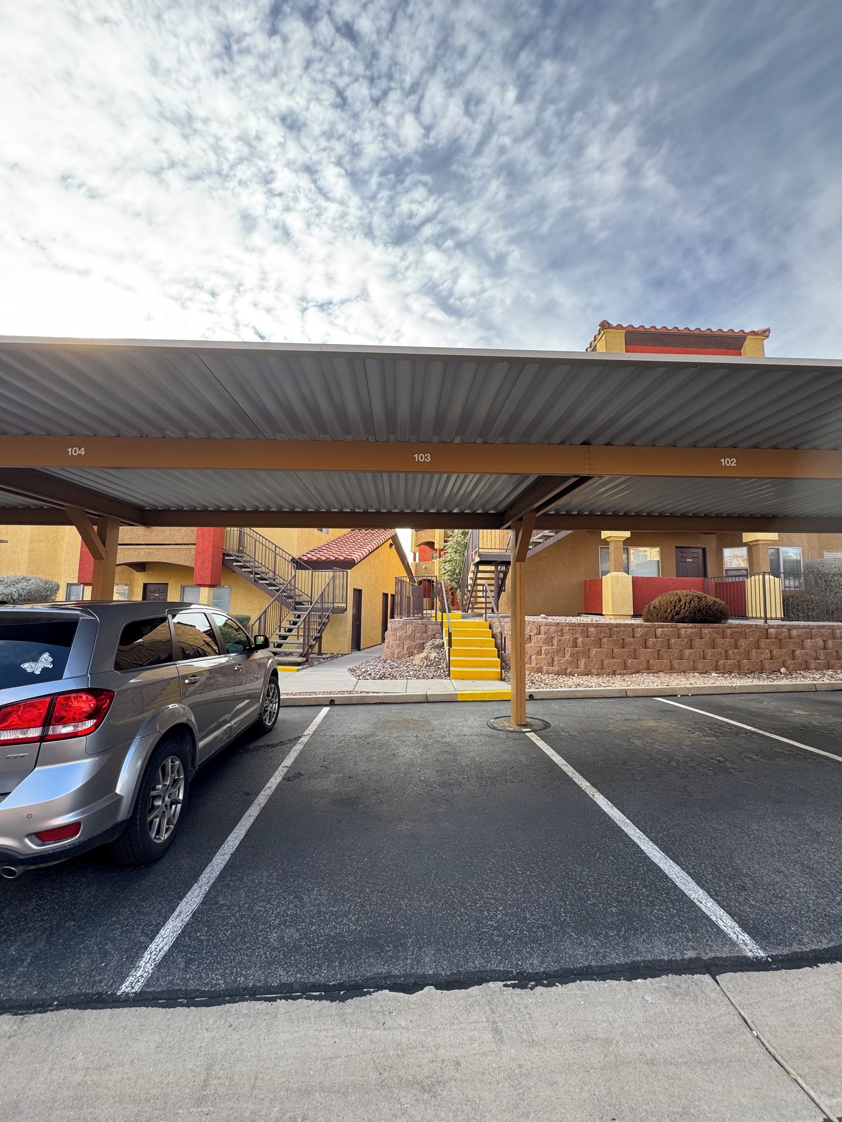 A parking area with a covered section, numbered 102 to 104, and a staircase leading to a building with a red and yellow facade.