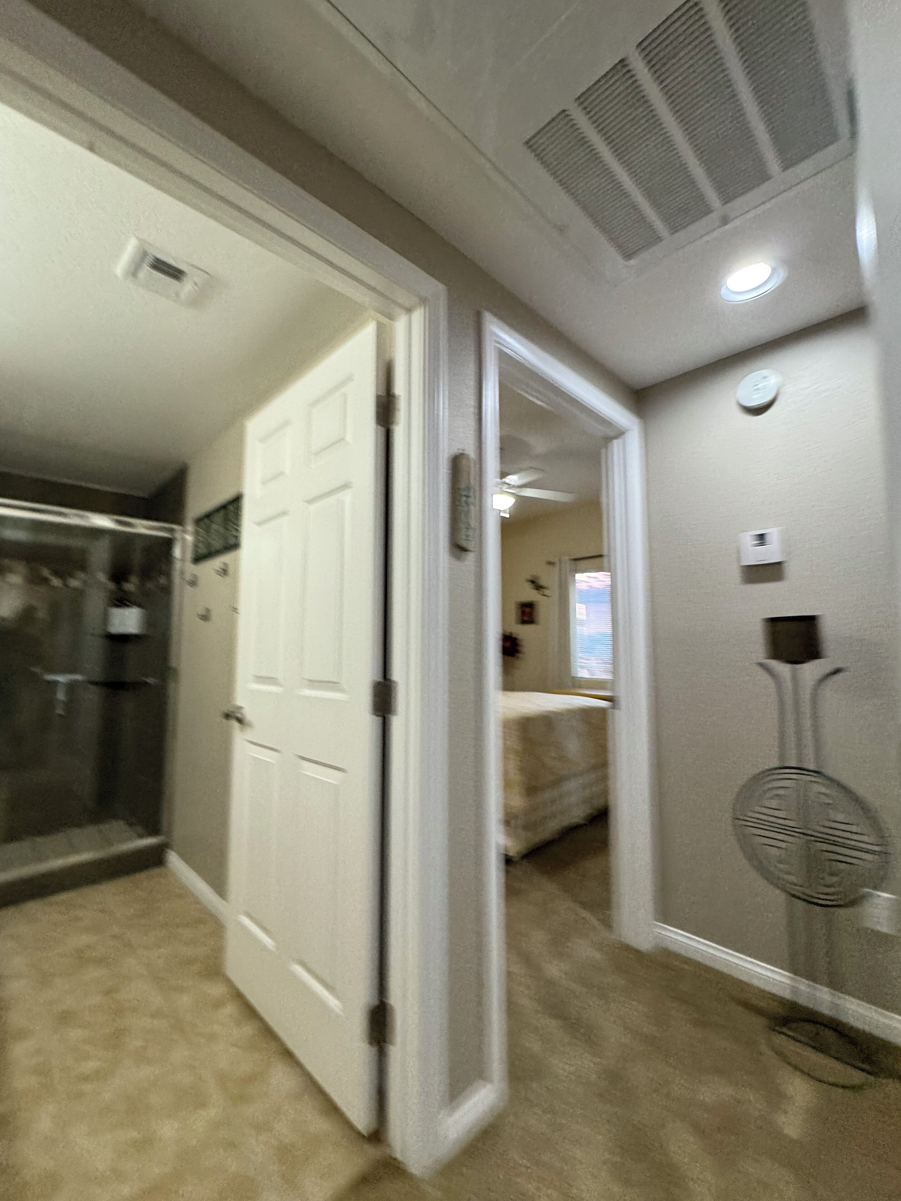 A hallway with a white door leading to a bathroom, a shower area, and a bedroom with a bed and a window.