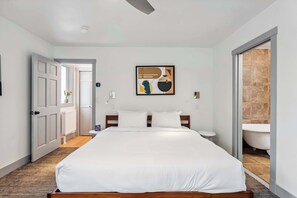 Un dormitorio con cama, un cuadro, un baño con bañera y una puerta que da a otra habitación.