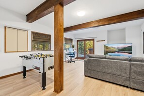 Un salon avec une table de baby-foot, un canapé gris et une grande fenêtre donnant sur les arbres.