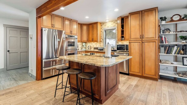 A kitchen with wooden cabinets, stainless steel appliances, a central island with stools, and a bookshelf with books and decorative items. - Breckenridge Mountain Retreat | Hot Tub & Views (Breckenridge)