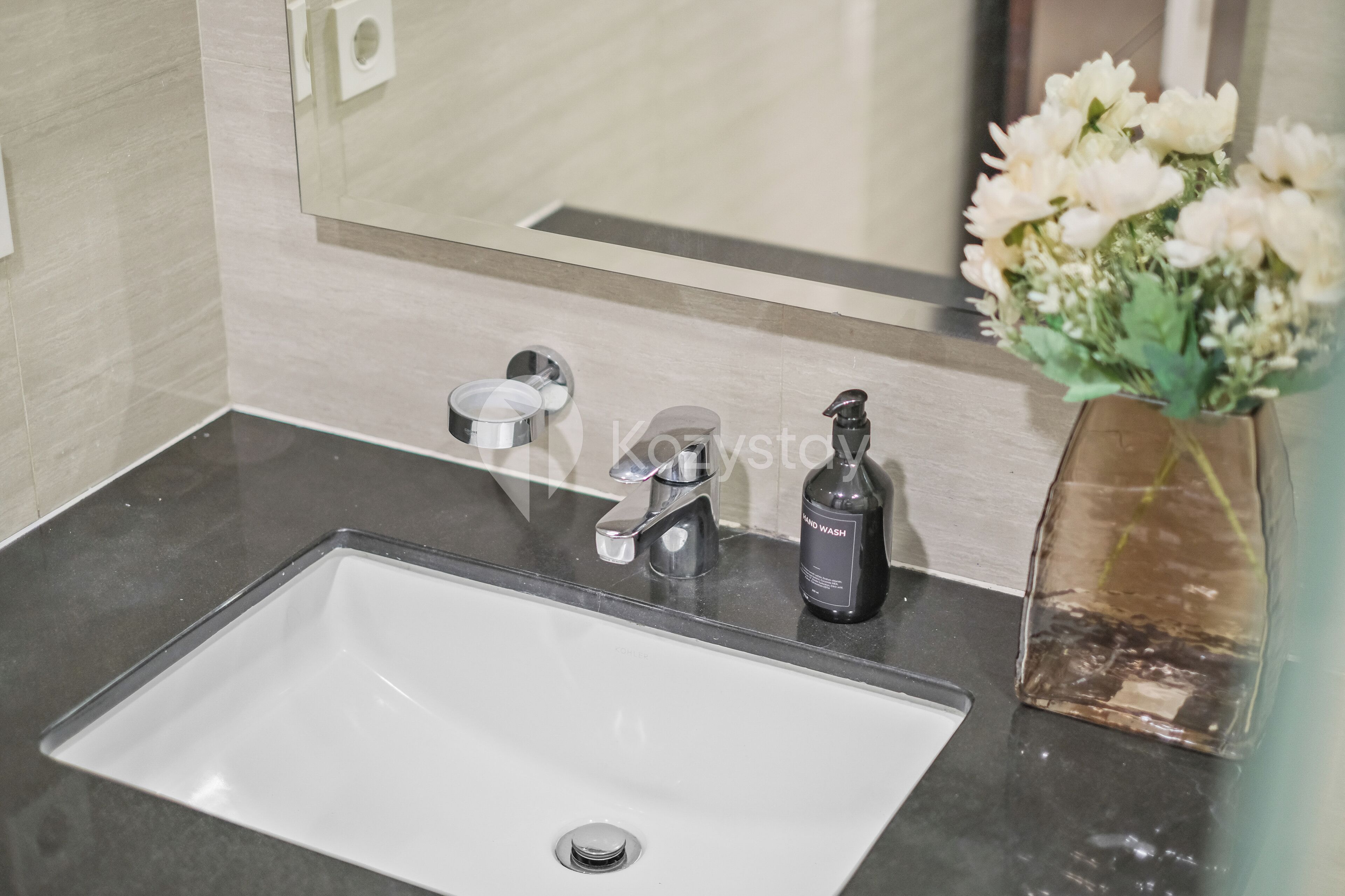 Une salle de bain avec un lavabo blanc, un miroir, un distributeur de savon noir et un vase rempli de fleurs blanches.