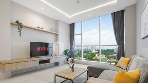 Una sala de estar moderna con un televisor grande mostrando 'NETFLIX', un sofá con cojines amarillos, una mesa de centro de madera y vistas de un paisaje urbano a través de ventanas amplias.