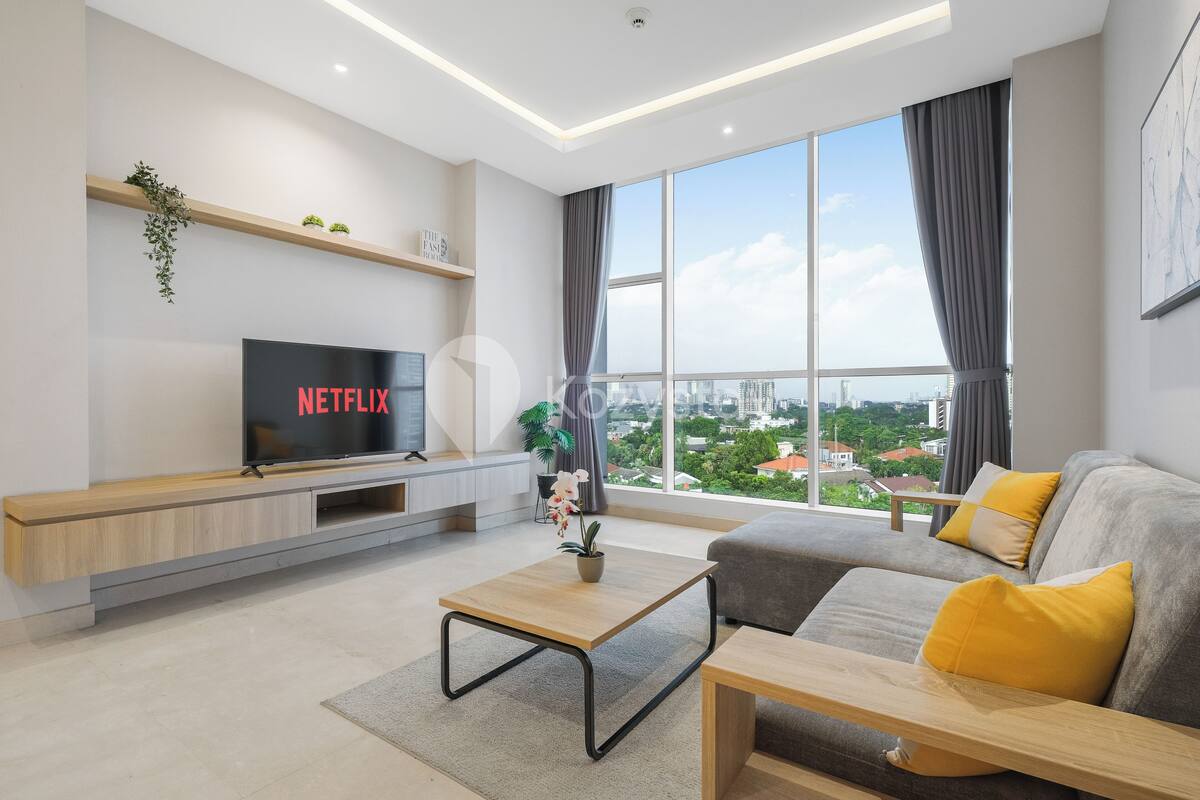 Una sala de estar moderna con un televisor grande mostrando 'NETFLIX', un sofá con cojines amarillos, una mesa de centro de madera y vistas de un paisaje urbano a través de ventanas amplias.