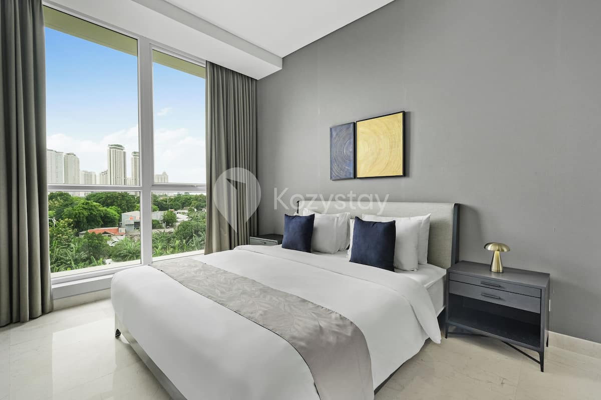 Un dormitorio moderno con una cama grande, una mesita de noche y vistas a un paisaje urbano a través de amplias ventanas.