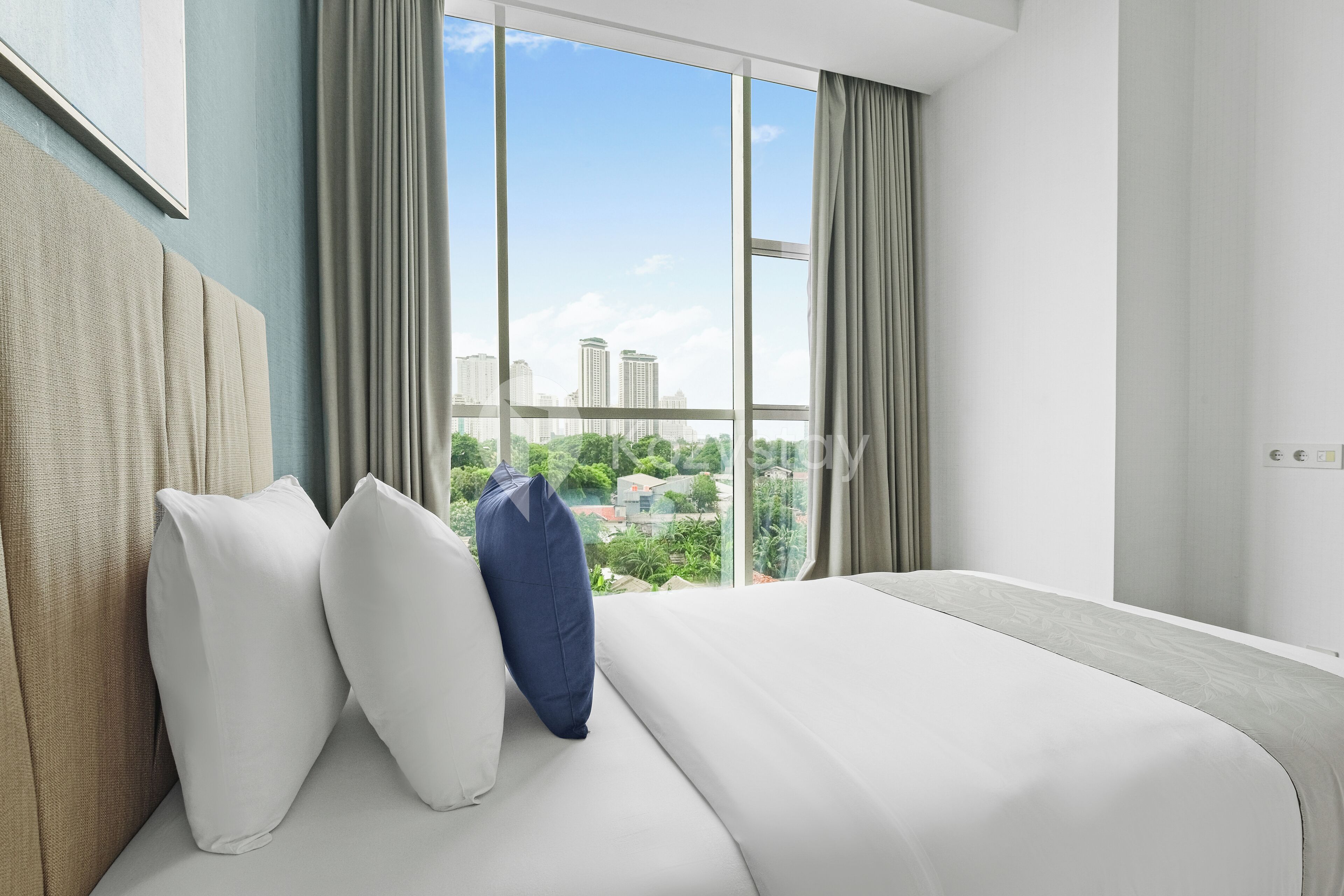 A modern hotel room with a large bed, two pillows, and a view of a cityscape through large windows.