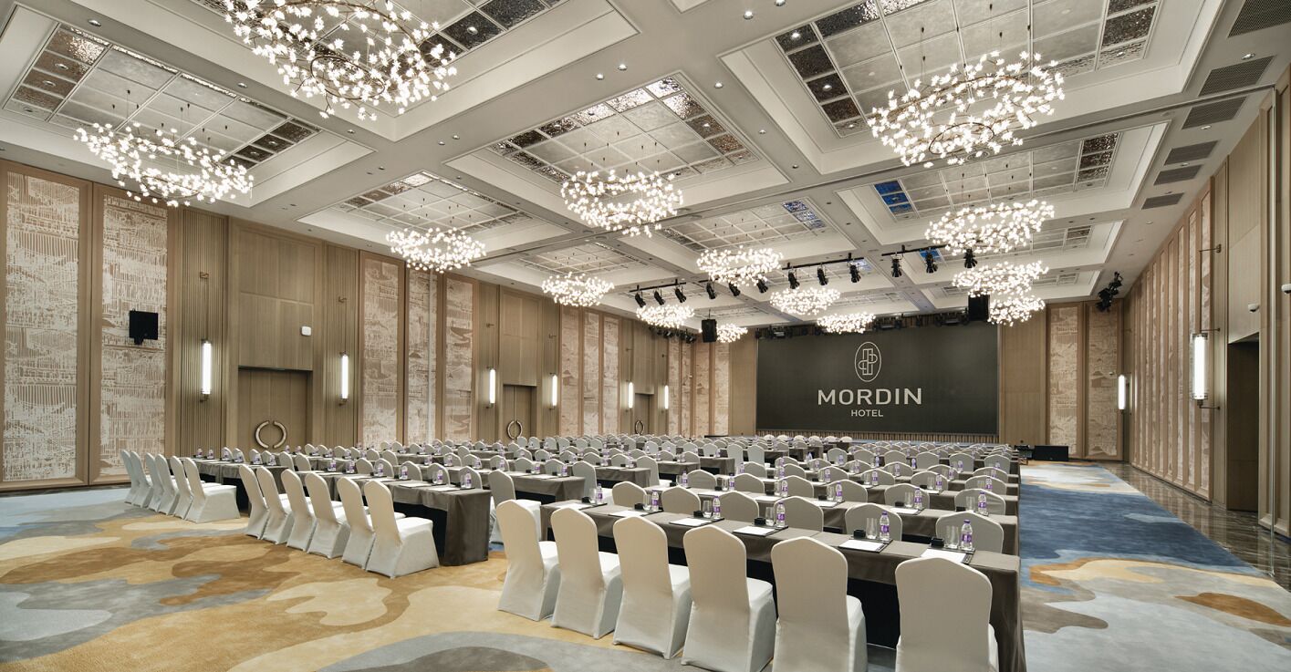 Uma sala de conferências ampla, com fileiras de mesas e cadeiras, um palco e um letreiro 'MORDIN HOTEL' na parede do fundo.