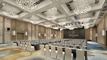 Uma sala de conferências ampla, com fileiras de mesas e cadeiras, um palco e um letreiro 'MORDIN HOTEL' na parede do fundo.