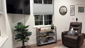 A living room with a brown leather chair, a small table with a plant, a bookshelf, and a large window with blinds.
