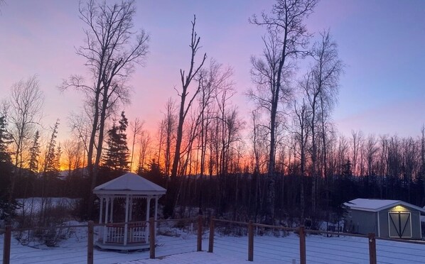 A gazebo and a small cabin in a snowy forest at sunset.
